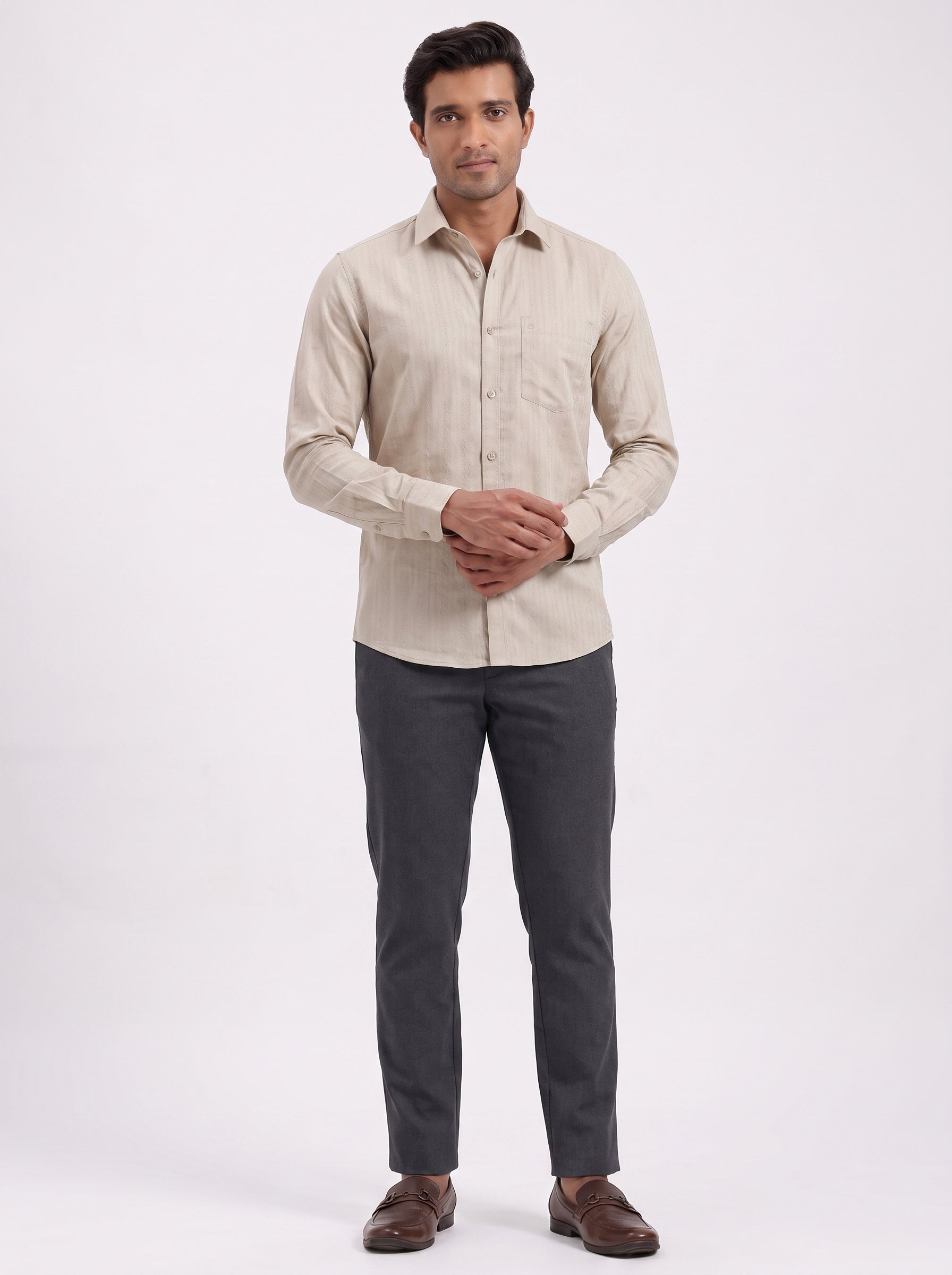 A man modeling a light beige long-sleeve linen shirt paired with dark gray trousers and brown leather shoes, standing against a plain white studio background.