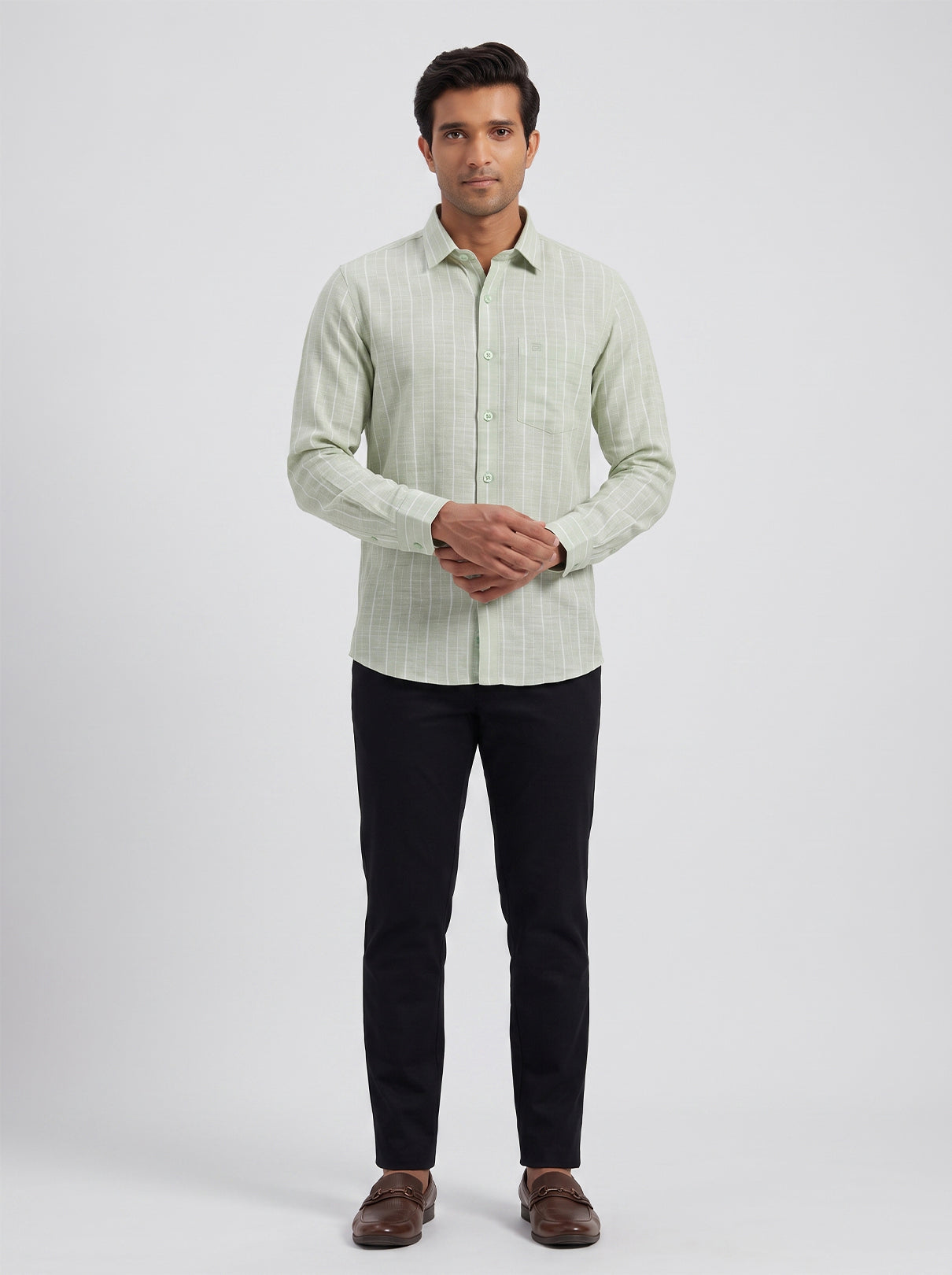 A man modeling a light sage green and white vertically striped long-sleeve button-down shirt, paired with black trousers and brown loafers, against a light gray studio background.