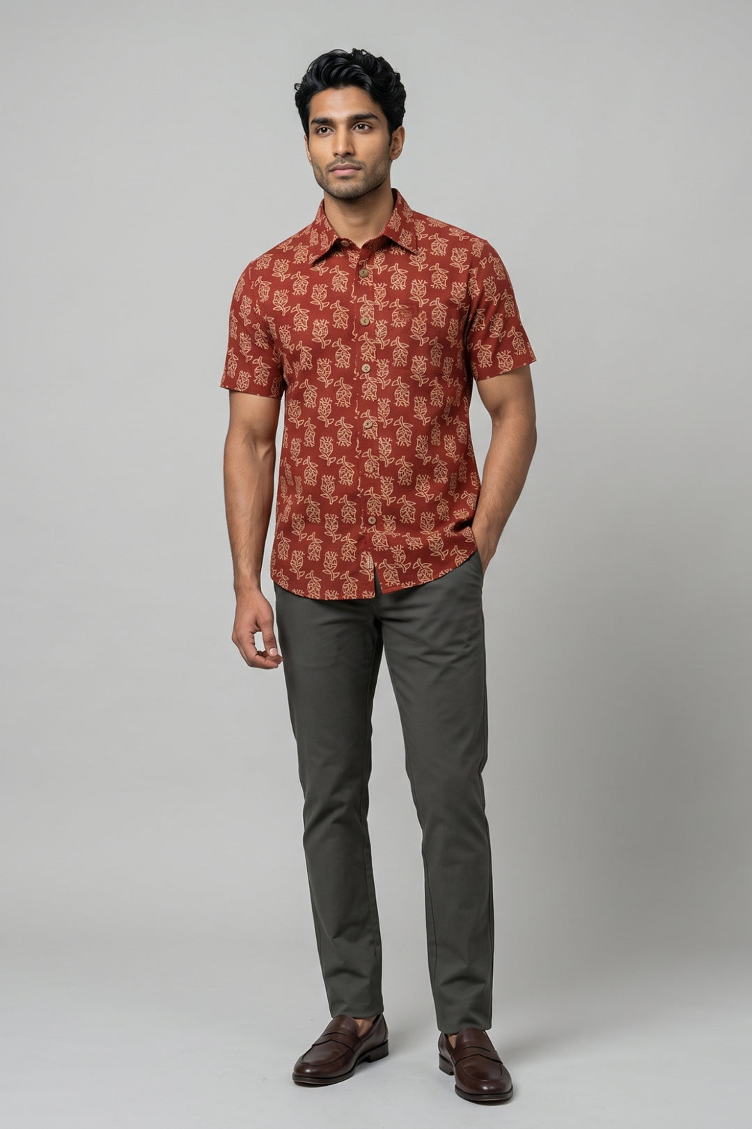 A man modeling a short-sleeve button-up shirt with a rust-red and brown traditional pattern, paired with dark green trousers and brown leather shoes, against a solid gray studio background.