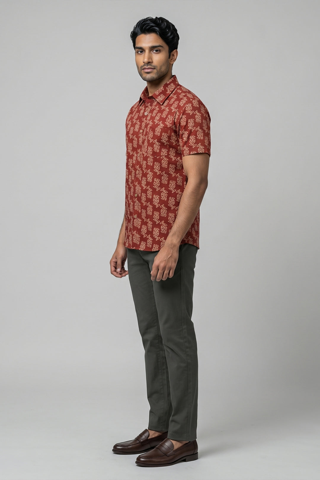 A man modeling a short-sleeve button-up shirt with a red and beige pattern, paired with dark gray trousers and brown leather shoes, against a solid green studio background.