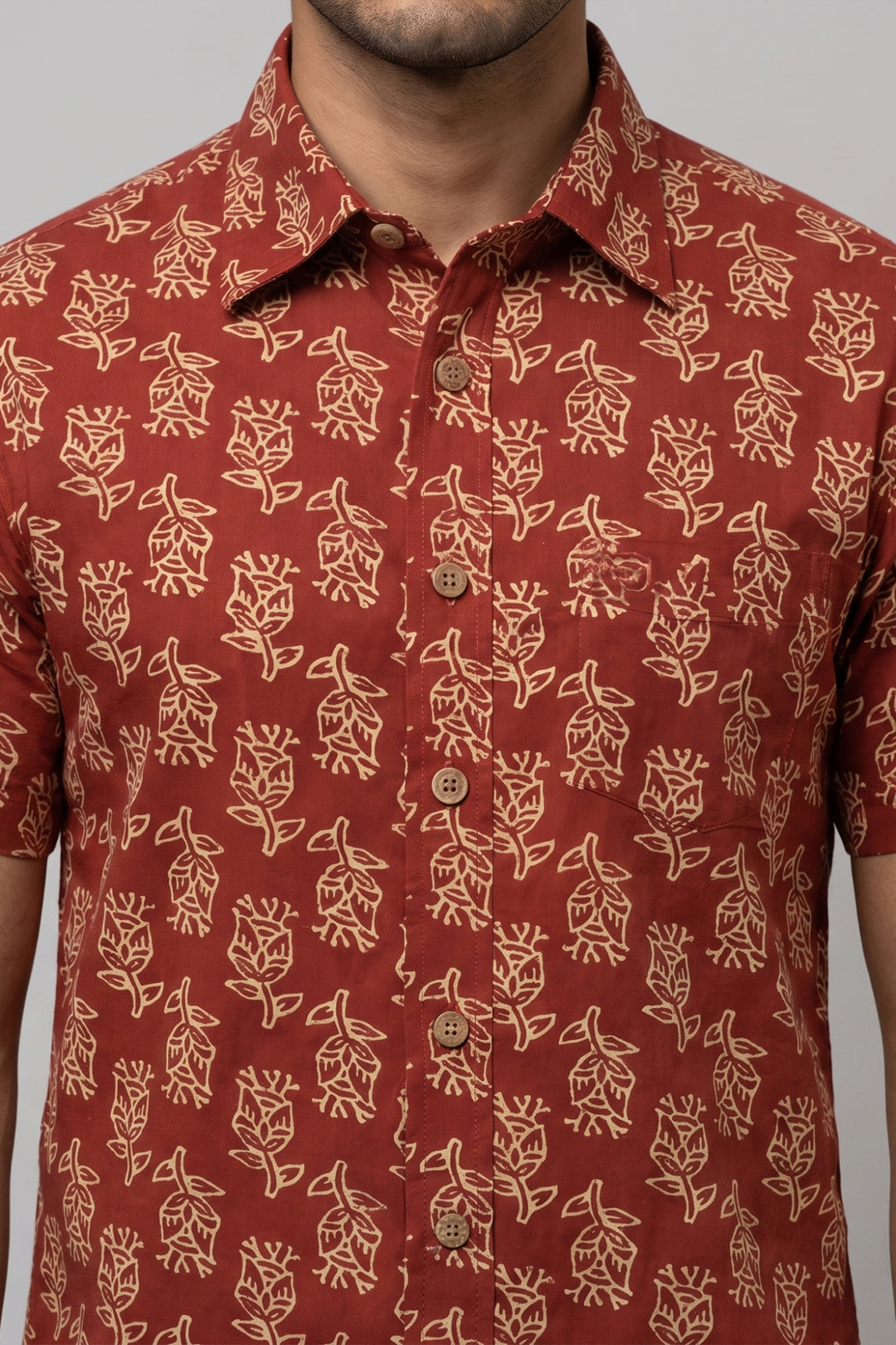A close-up of a man wearing a short-sleeve button-up shirt in rust red with a beige block-printed floral pattern. The shirt features brown buttons and is shown against a neutral gray background.