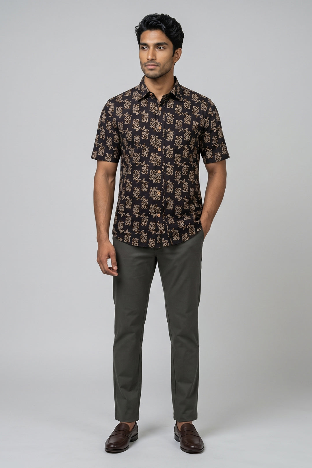 A man modeling a short-sleeve button-up shirt with a dark Black and beige floral pattern, paired with dark green trousers and brown leather shoes, against a solid light gray background.