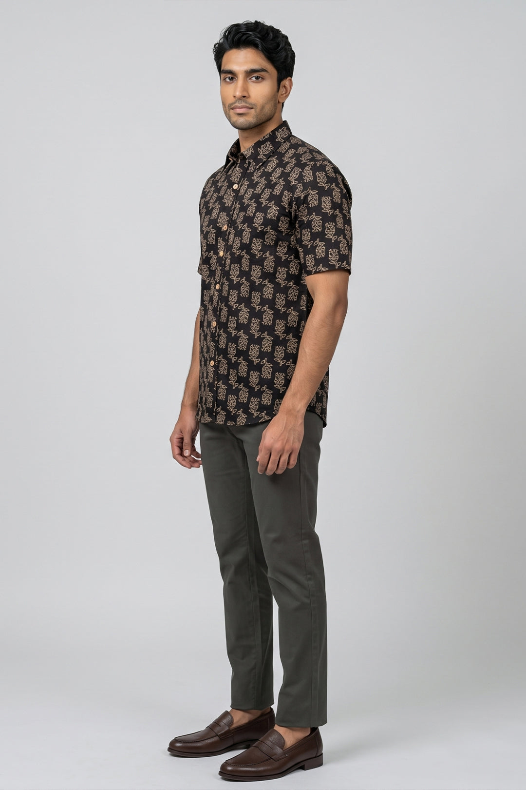 A man modeling a short-sleeve button-up shirt with a brown and beige floral pattern, paired with dark olive trousers and brown leather loafers, against a light gray studio background.