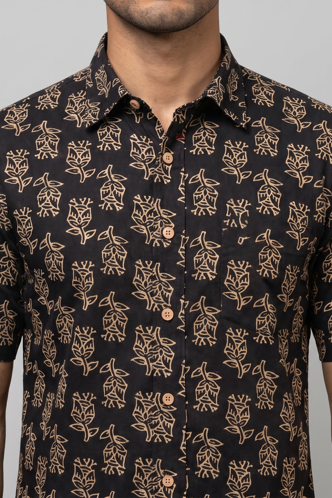 A man wearing a short-sleeve button-up shirt with a black base and a tan floral block print pattern, shown from the chest up against a neutral gray background.