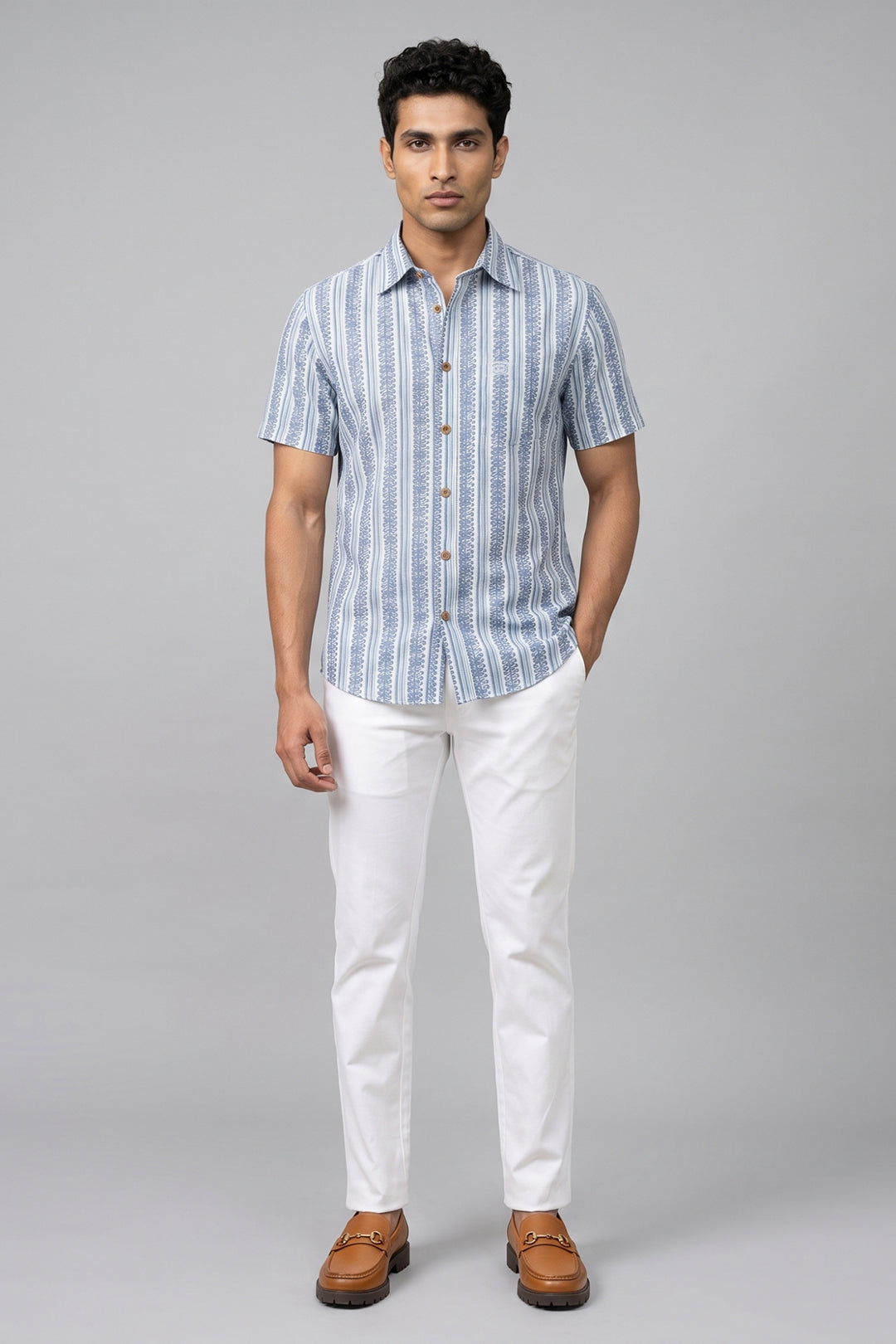 A man modeling a short-sleeve button-up shirt with vertical blue and white stripes, paired with white trousers and brown leather loafers, against a solid gray studio background.