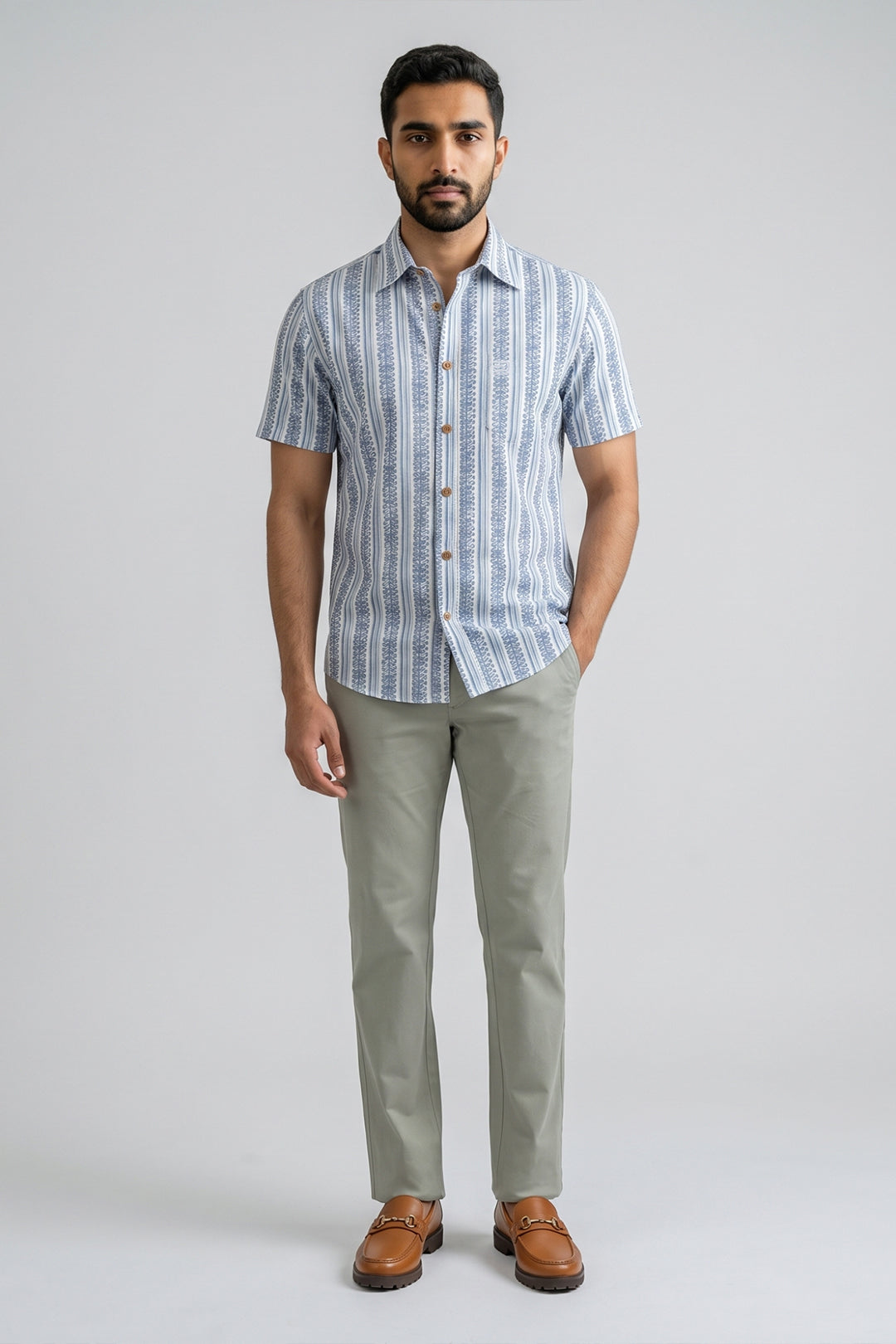 A man modeling a short-sleeve button-up shirt with vertical blue and white stripes, paired with light olive green trousers and brown leather loafers, against a solid light gray background.