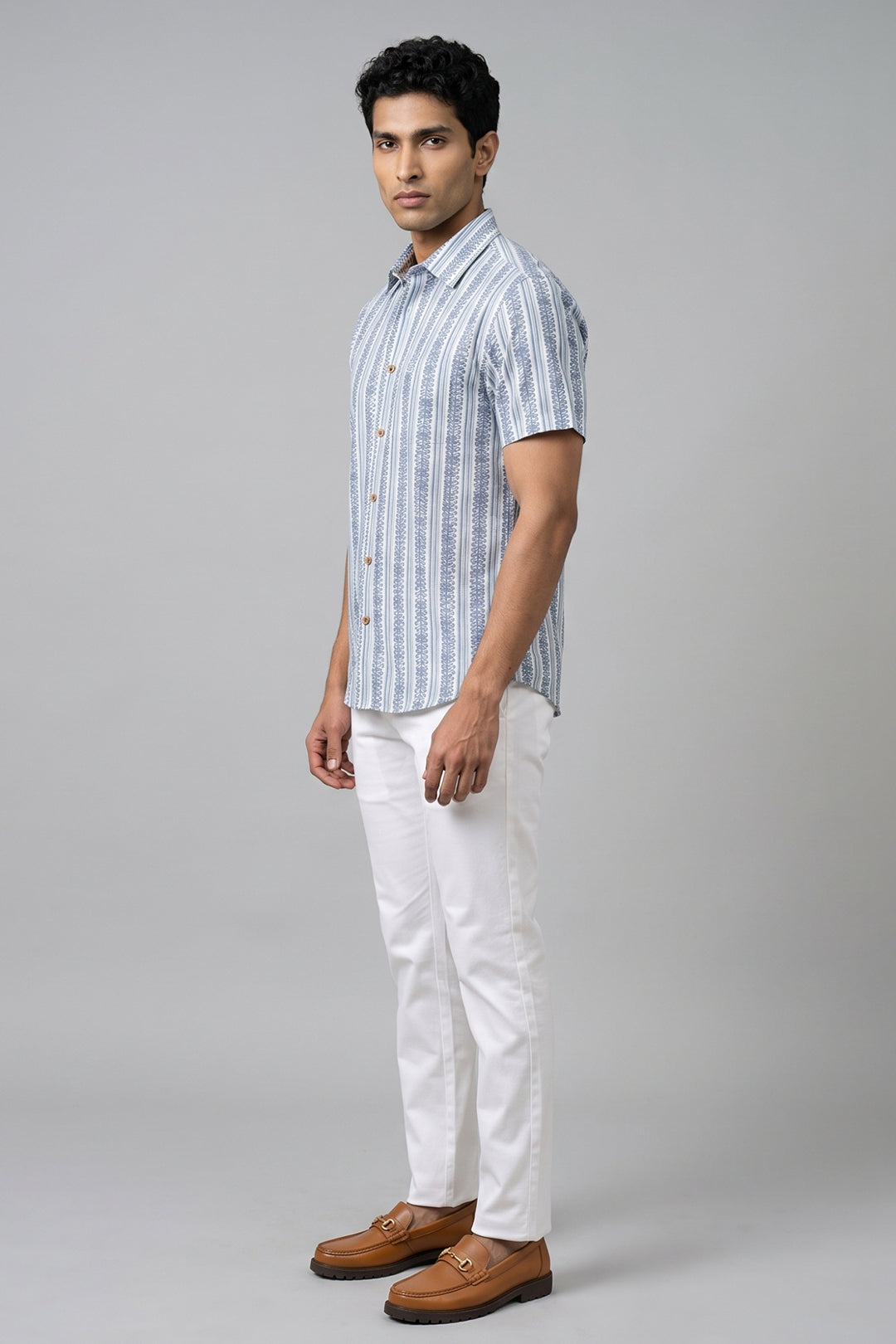 A man modeling a short-sleeve button-up shirt with vertical blue and white stripes, paired with white trousers and brown loafers, against a solid gray studio background.
