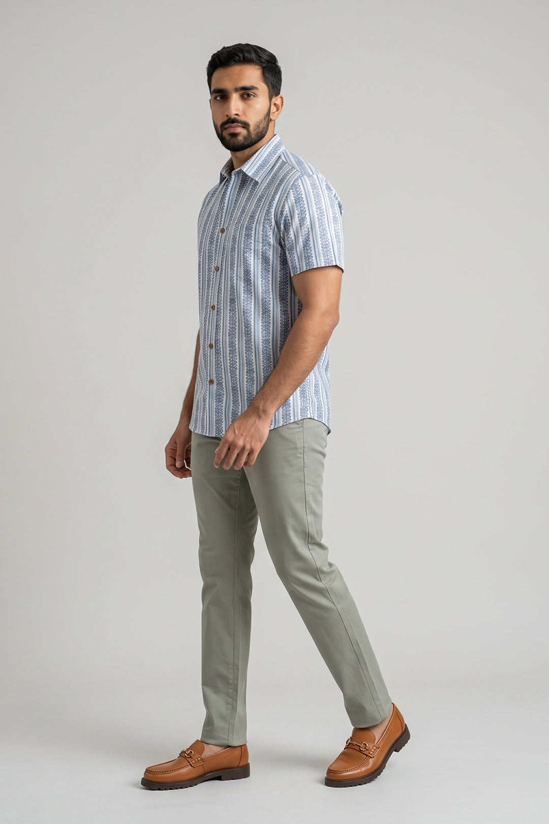 A man wearing a light blue and white vertically striped short-sleeve button-up shirt, olive green trousers, and brown leather loafers, standing against a solid light gray studio background.