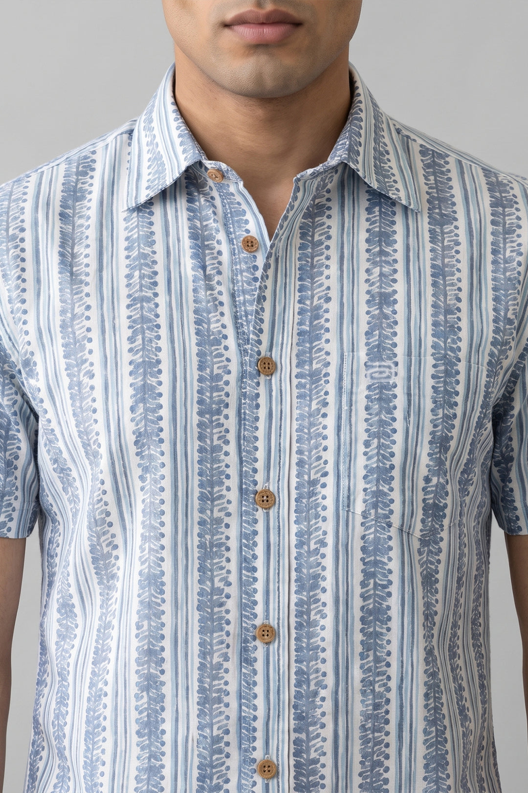 A close-up of a man wearing a short-sleeve button-up shirt with vertical blue and white leafy patterns and brown buttons, shown against a plain light gray background.
