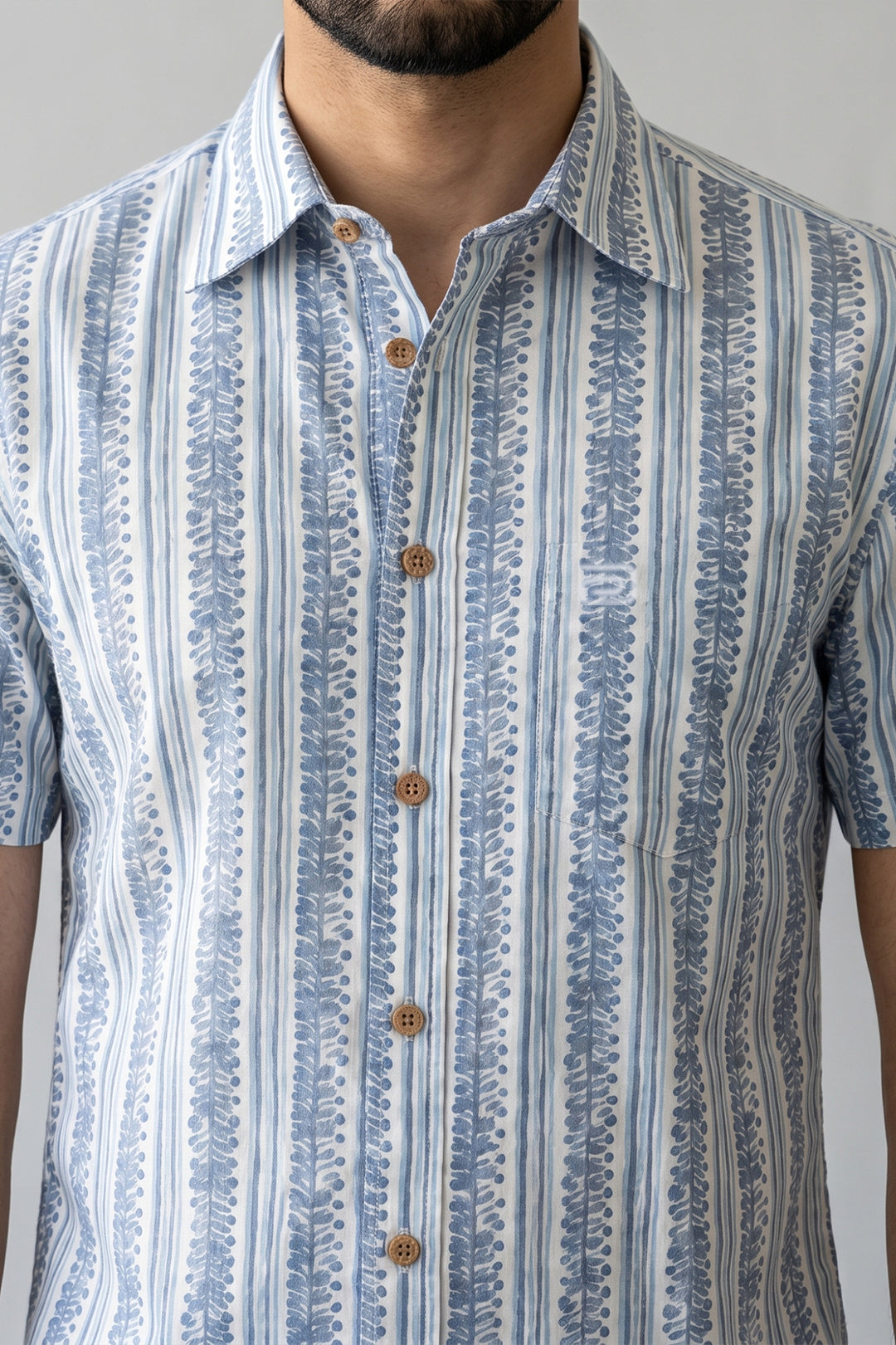 A close-up of a man wearing a short-sleeve button-up shirt with vertical blue and white patterns, featuring brown buttons.