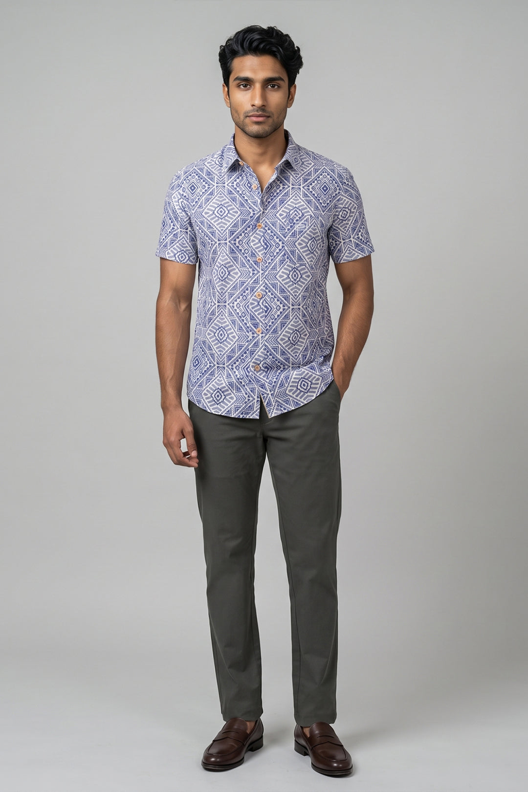 A man modeling a short-sleeve button-up shirt with a blue and white geometric pattern, paired with dark green trousers and brown leather shoes, against a solid gray studio background.