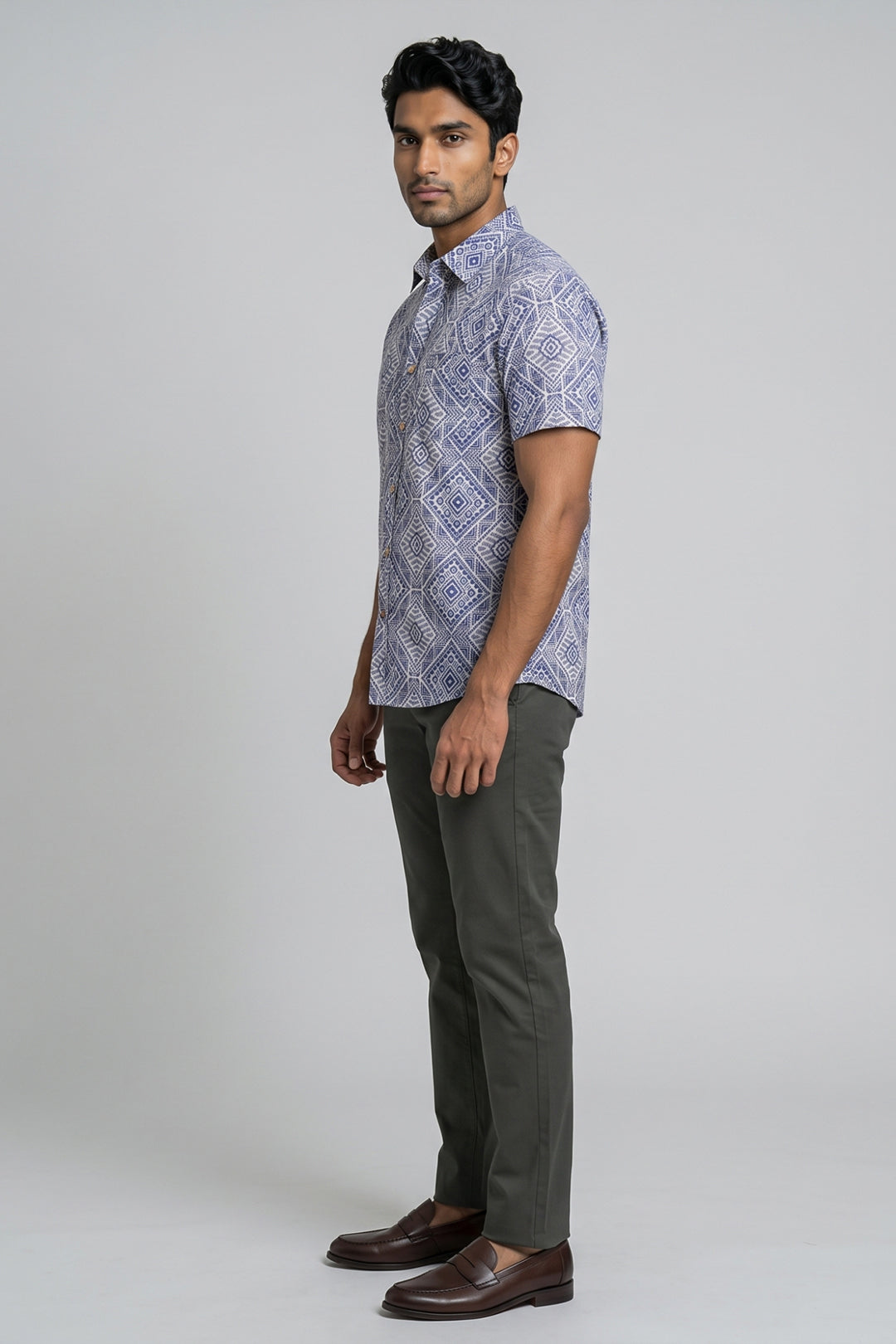 A man modeling a short-sleeve button-up shirt with a blue and white geometric pattern, paired with dark olive trousers and brown leather shoes, against a plain light gray studio background.