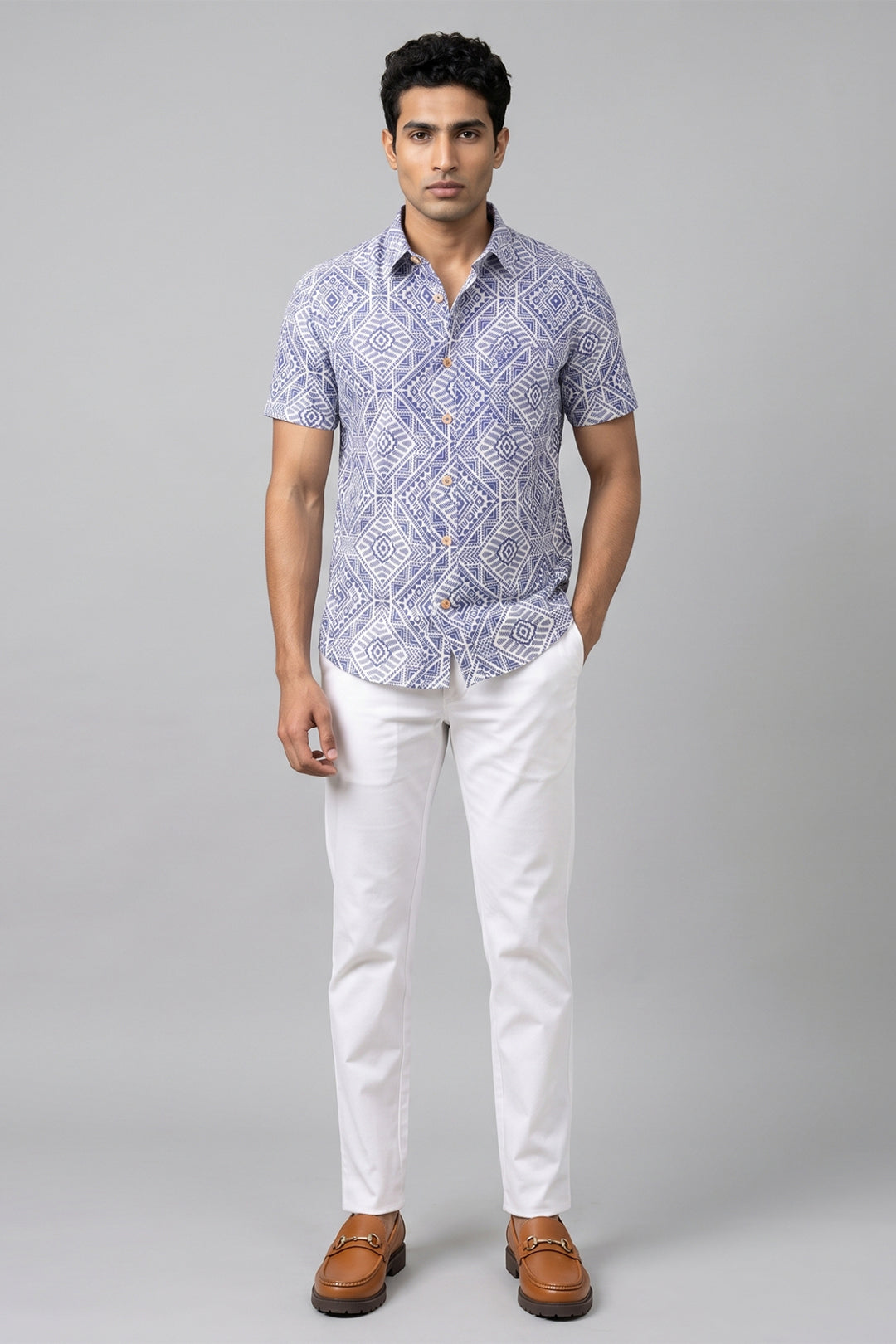 A man modeling a short-sleeve button-up shirt with a blue and white geometric pattern, paired with white trousers and brown leather loafers, against a solid gray studio background.