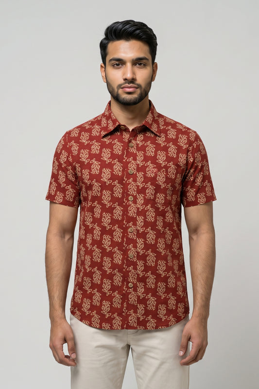 A man wearing a short-sleeve button-up shirt with a rust red and beige floral pattern, paired with light beige trousers, standing against a solid light gray background.
