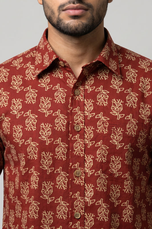 A close-up of a man wearing a rust-red button-down shirt with a beige floral block print pattern. The image is cropped from the lower face to the chest, showing the collar, front buttons, and fabric texture against a neutral gray background.