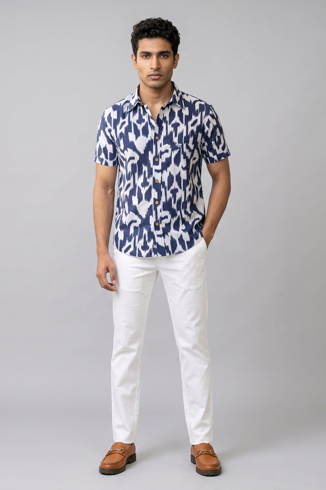 A man modeling a short-sleeve button-up shirt with a navy blue and white ikat pattern, paired with white trousers and brown leather loafers, against a solid gray studio background.