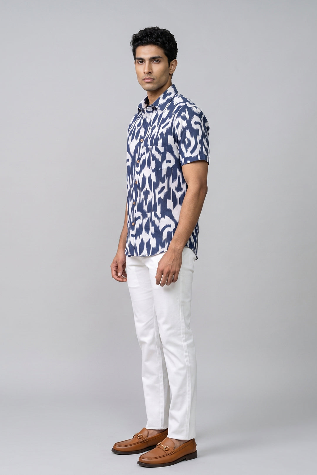 A man modeling a short-sleeve button-up shirt with a navy and white abstract ikat pattern, paired with white trousers and brown leather loafers, against a solid light gray background.