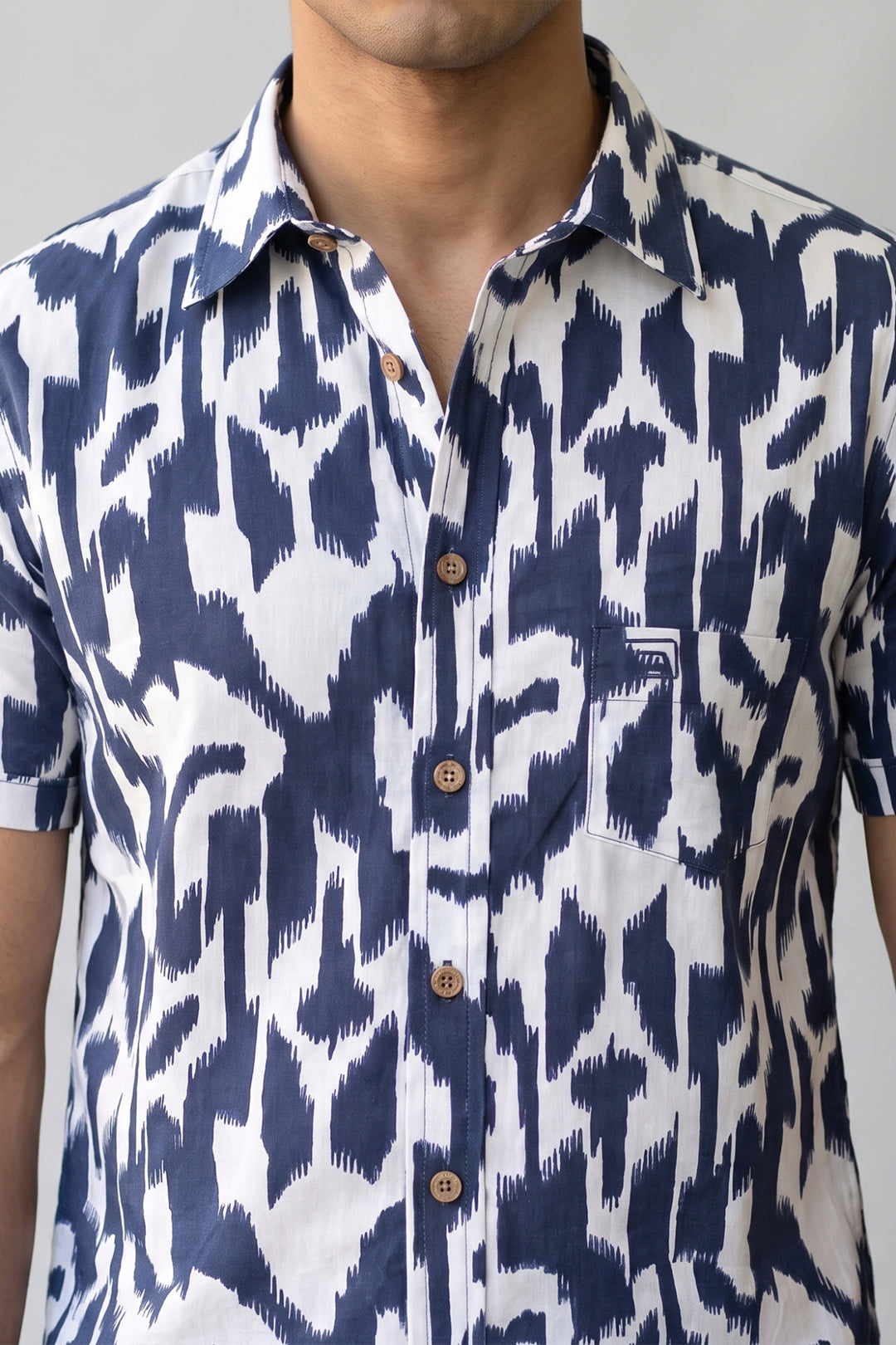 A close-up of a man wearing a short-sleeve button-up shirt with a navy blue and white ikat pattern. The shirt features brown buttons and a small chest pocket with a visible 'FILA' brand label.