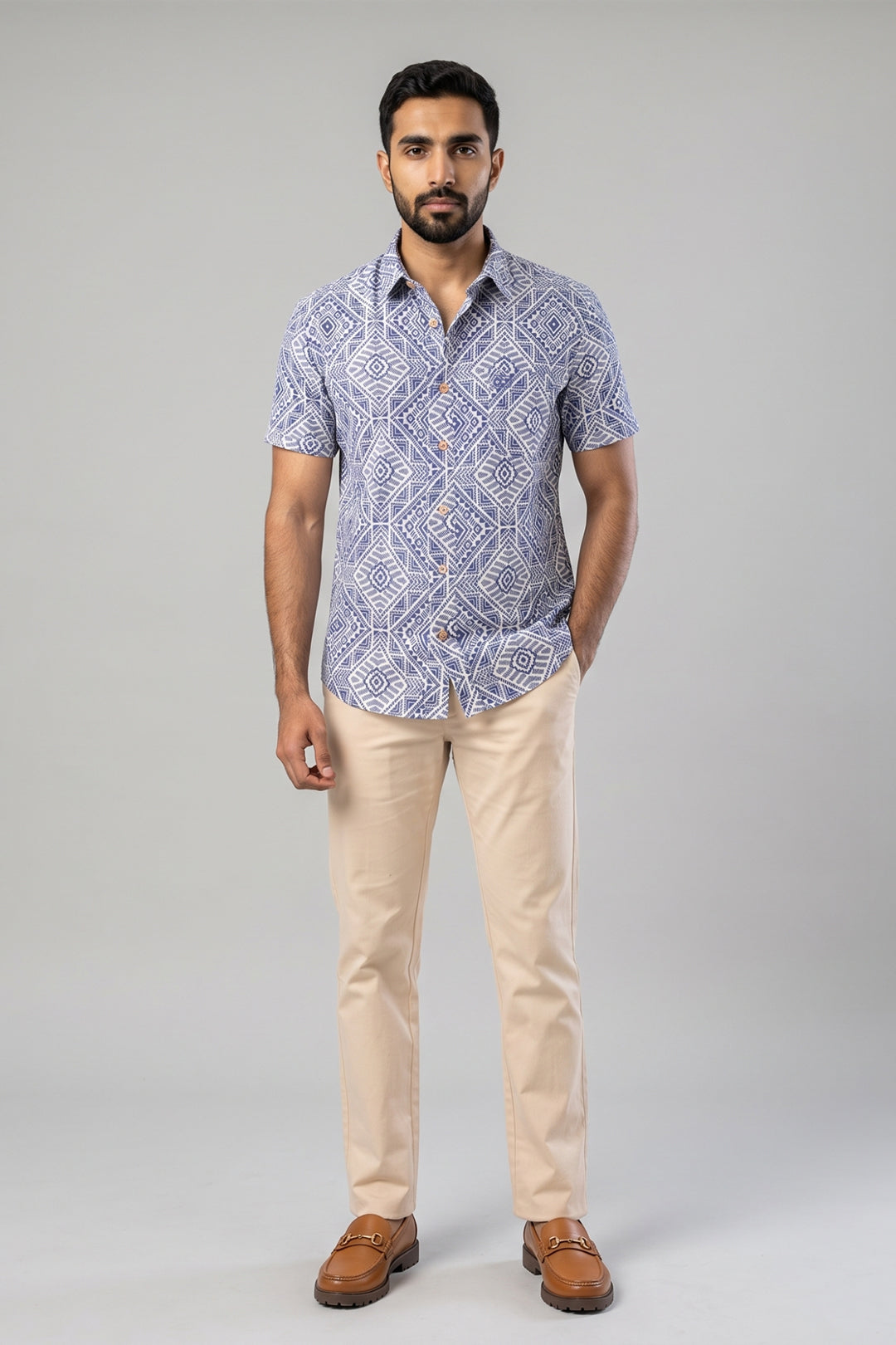 A man modeling a short-sleeve button-up shirt with a blue and white geometric pattern, paired with beige trousers and brown leather loafers, against a solid gray studio background.