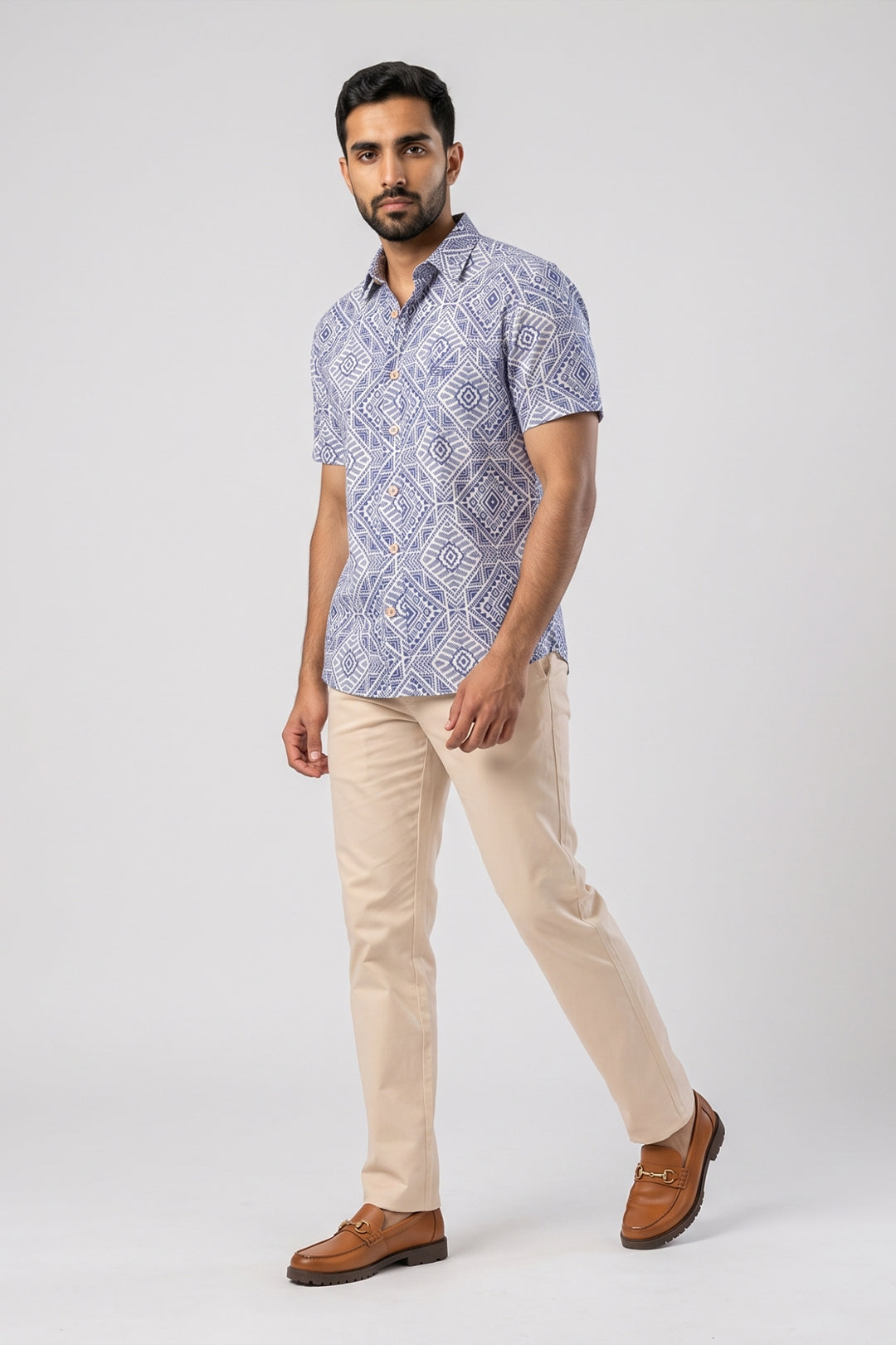 A man modeling a short-sleeve button-up shirt with a blue and white geometric pattern, paired with beige trousers and brown loafers, against a light gray studio background.