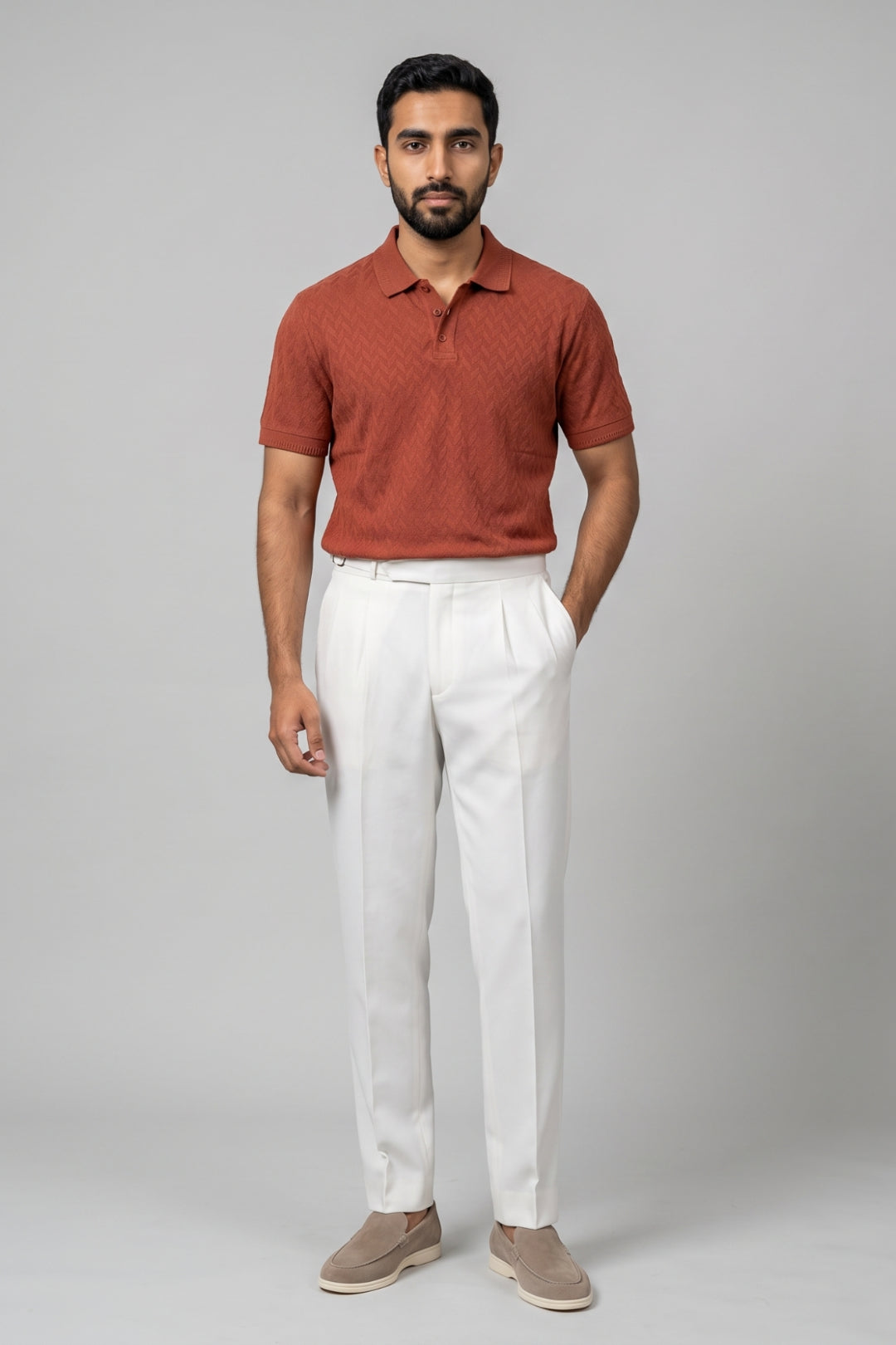 A man modeling a short-sleeve polo shirt in rust orange with a subtle textured pattern, paired with white trousers and beige slip-on shoes, against a solid gray studio background.