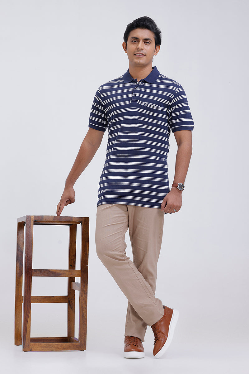 Man wearing a striped polo shirt and beige pants standing next to a wooden stool on a white background