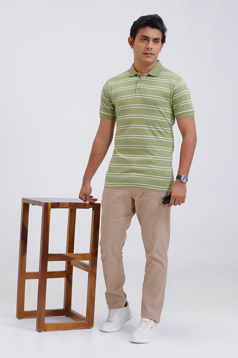 Man wearing a green striped polo shirt and beige pants standing next to a wooden stool on a white background