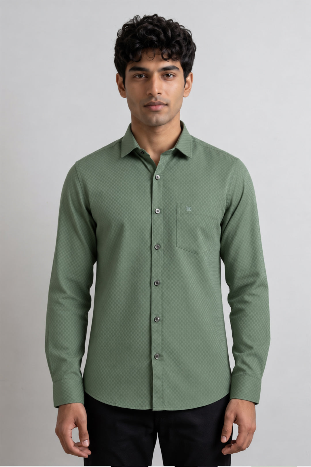 A man wearing a green long-sleeve button-down shirt with a subtle textured pattern, paired with black trousers, standing against a light gray studio background.