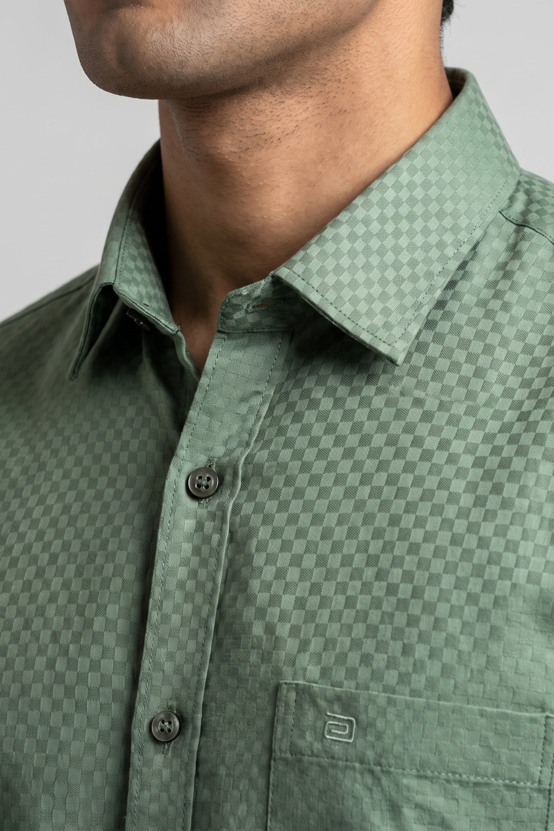 Close-up of a man wearing a green checkered button-down shirt, showing the collar, chest, and buttons against a neutral gray background.