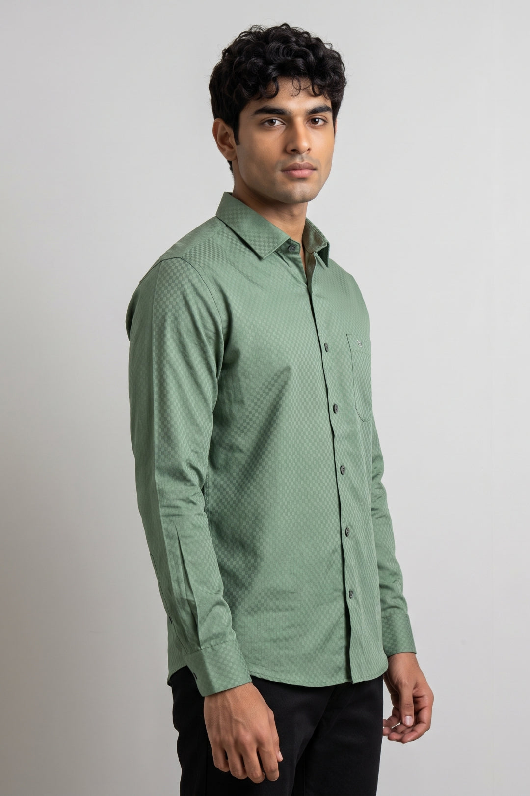 A man wearing a green long-sleeve button-up shirt with a subtle textured pattern, paired with black trousers, standing against a plain light gray studio background.